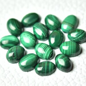 May include: A group of 14 oval-shaped malachite gemstones, each with a unique green and white pattern. The stones are arranged in a circle on a white surface.