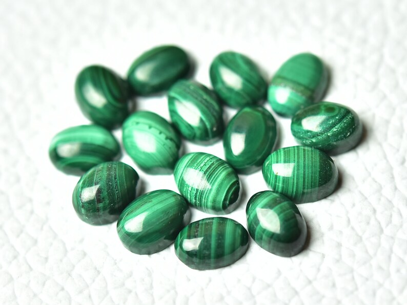 May include: A group of 14 oval-shaped malachite gemstones, each with a unique green and white pattern. The stones are arranged in a circle on a white surface.