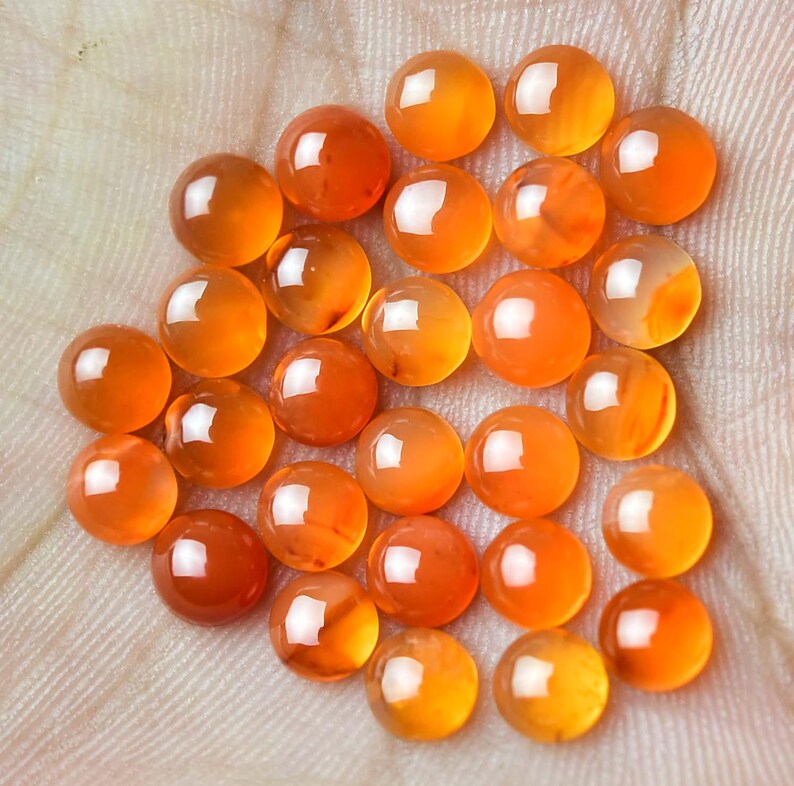 Pu&ograve; includere: Una collezione di gemme rotonde e lucide nei toni dell'arancione e dell'ambra. Le gemme sono lisce e riflettenti, con una finitura lucida. La disposizione delle pietre suggerisce che sono sciolte, forse per la creazione di gioielli o la collezione.