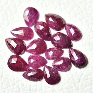 Pode incluir: Uma coleção de rubis facetados em forma de pêra, em vários tamanhos. As pedras preciosas exibem uma cor vermelha profunda e rica com múltiplas facetas que capturam a luz. Os rubis estão espalhados sobre uma superfície branca.