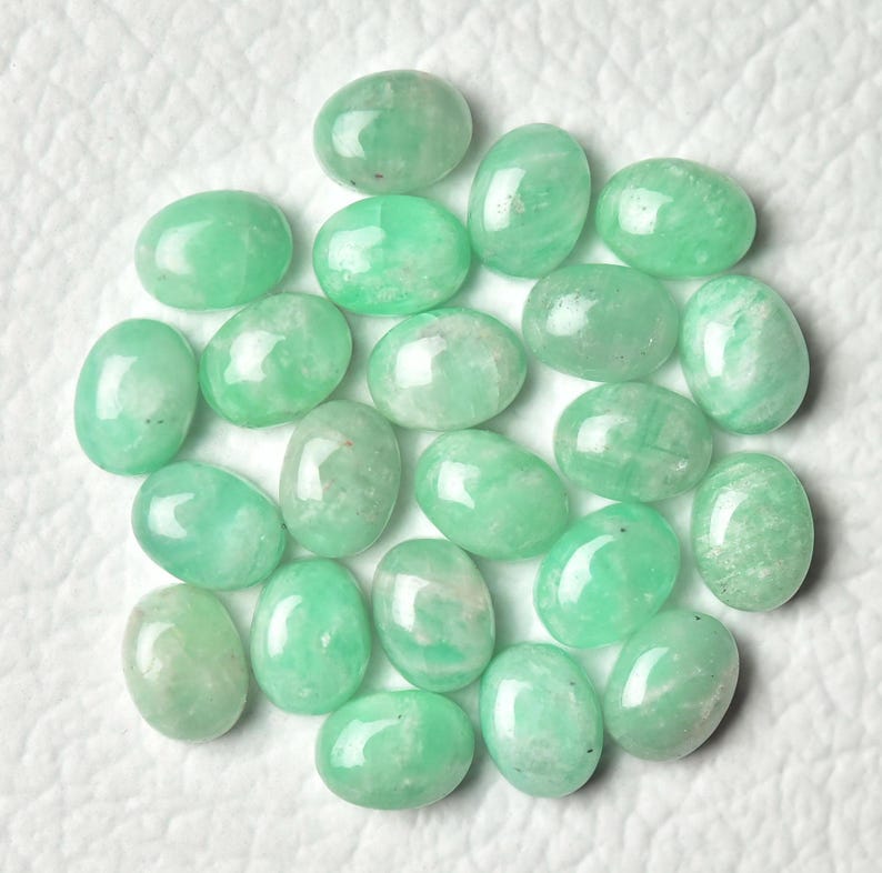 Peut inclure: Une collection de 18 pierres pr&eacute;cieuses vertes de forme ovale, probablement de l'amazonite, dispos&eacute;es en cercle sur une surface blanche.