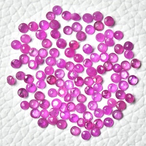 May include: A collection of approximately 100 pink ruby gemstones, each with a faceted round shape. The gemstones are scattered on a white surface.