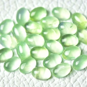 May include: A collection of oval-shaped, translucent green gemstones. The stones have a smooth, polished finish and are arranged in a loose pile.