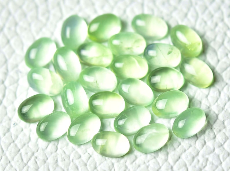 May include: A collection of oval-shaped, translucent green gemstones. The stones have a smooth, polished finish and are arranged in a loose pile.