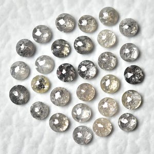 May include: A collection of round, faceted diamonds in various shades of gray, silver, and yellow. The gemstones are scattered on a white surface, showcasing their unique textures and brilliance. These diamonds are ideal for jewelry making.