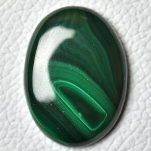 May include: A green malachite cabochon gemstone with a swirling pattern. The stone is oval shaped and has a smooth, polished surface.