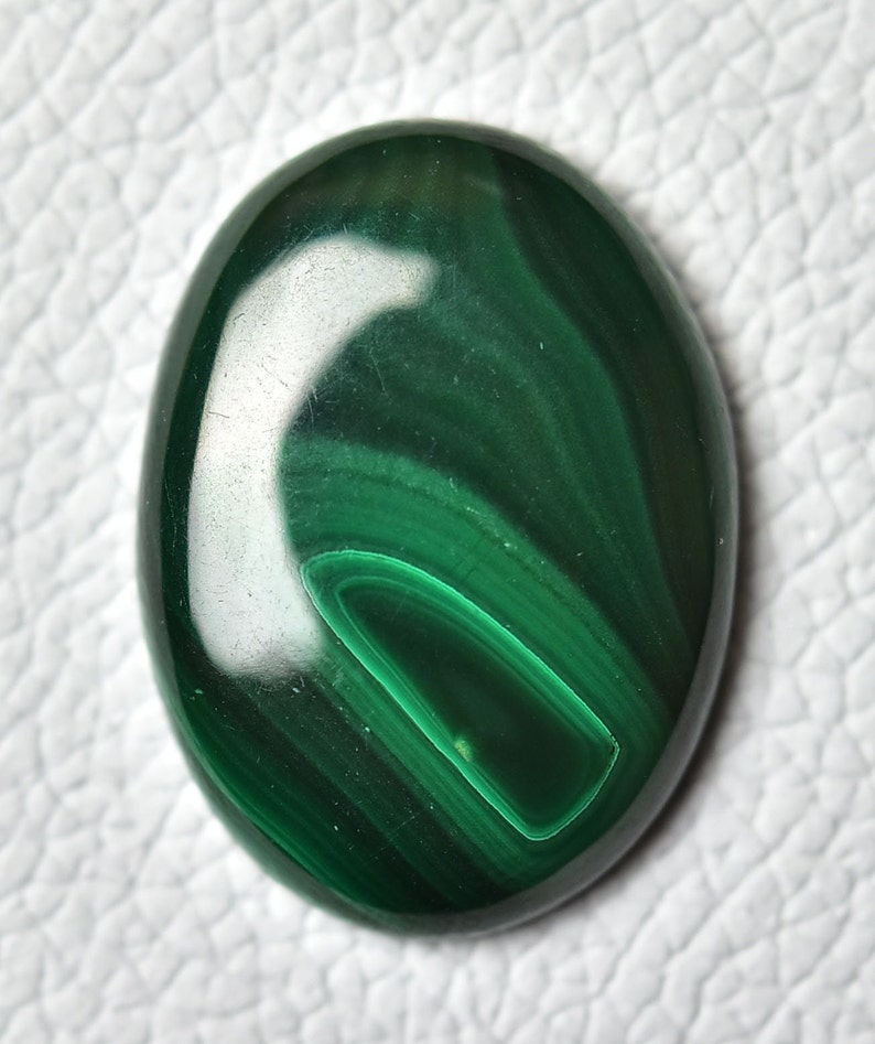 May include: A green malachite cabochon gemstone with a swirling pattern. The stone is oval shaped and has a smooth, polished surface.