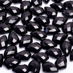 May include: A collection of black gemstone beads in a faceted teardrop shape. The beads are arranged in a random pattern on a white surface.