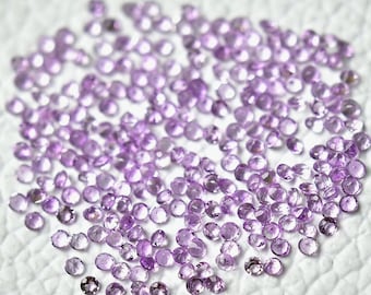 May include: A large group of small, round, light purple gemstones. The gemstones are scattered on a white surface.