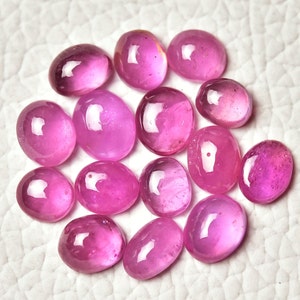May include: A collection of oval-shaped, polished pink gemstones. The stones vary slightly in size and shade, with a glossy, translucent appearance. The gemstones are arranged on a white surface, showcasing their vibrant colour and smooth texture.