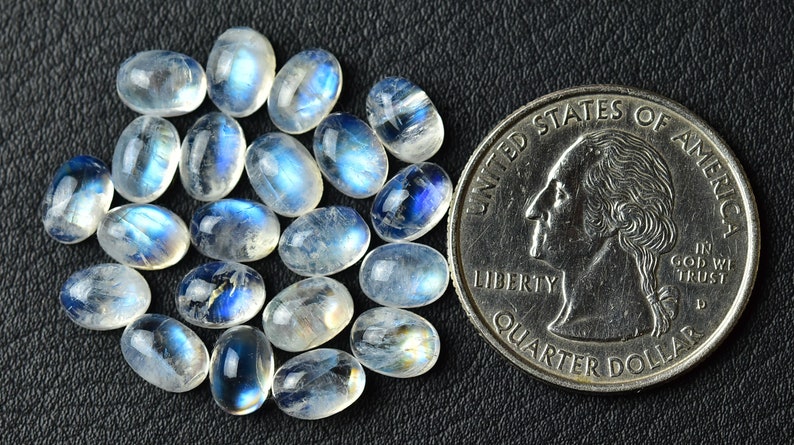 May include: A collection of oval-shaped moonstone cabochons, ranging in size from small to medium, with a milky white color and iridescent blue sheen. The stones are arranged in a loose pile and are compared to a US quarter for size reference.