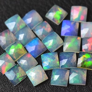 Può includere: Una collezione di 20 cabochon di opale iridescente, ciascuno tagliato a forma quadrata con una parte superiore sfaccettata. Gli opali sono di colore bianco latte con bagliori di verde, blu e rosso.