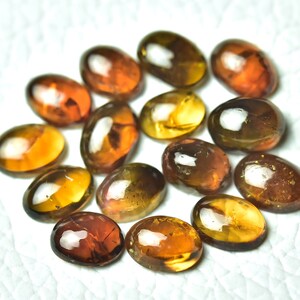 May include: A collection of 13 oval-shaped gemstones in shades of brown, yellow, and orange. The gemstones have a glossy finish and are arranged in a circular pattern.