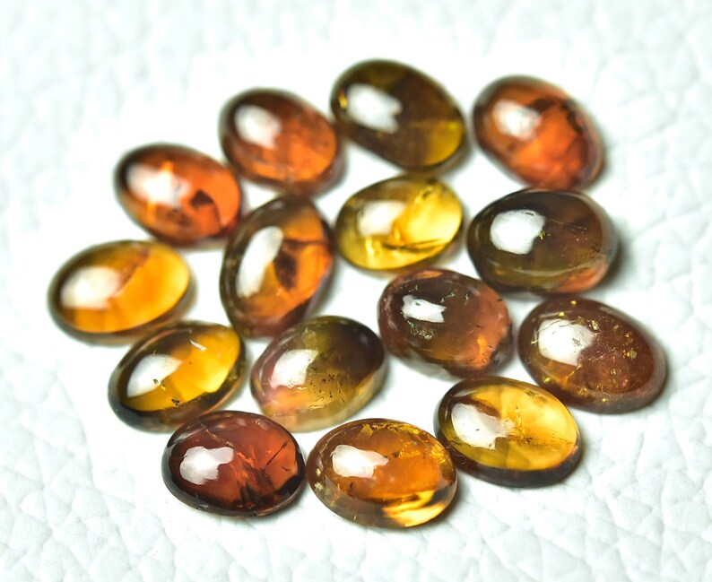 May include: A collection of 13 oval-shaped gemstones in shades of brown, yellow, and orange. The gemstones have a glossy finish and are arranged in a circular pattern.