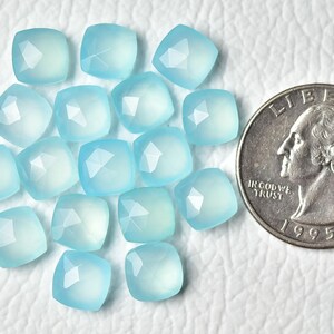 K&ouml;nnte beinhalten: Eine Sammlung von 15 hellblauen Chalcedon-Edelsteinkugeln, jede mit einer facettierten quadratischen Form. Die Kugeln sind in einer Gruppe auf einer wei&szlig;en Oberfl&auml;che angeordnet, mit einer US-amerikanischen 25-Cent-M&uuml;nze als Gr&ouml;&szlig;envergleich.