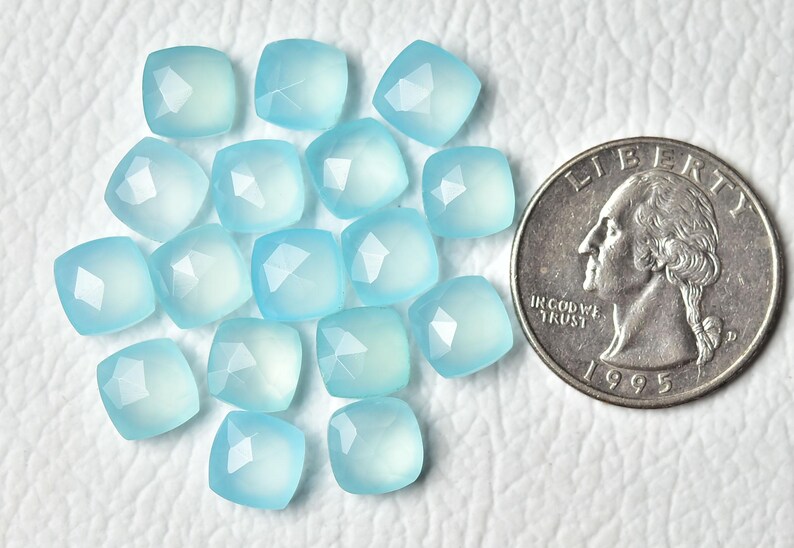K&ouml;nnte beinhalten: Eine Sammlung von 15 hellblauen Chalcedon-Edelsteinkugeln, jede mit einer facettierten quadratischen Form. Die Kugeln sind in einer Gruppe auf einer wei&szlig;en Oberfl&auml;che angeordnet, mit einer US-amerikanischen 25-Cent-M&uuml;nze als Gr&ouml;&szlig;envergleich.