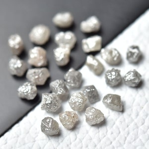 Puede incluir: Una colecci&oacute;n de diamantes grises en bruto y sin pulir. Los diamantes son de varios tama&ntilde;os y formas, y est&aacute;n esparcidos sobre una superficie blanca.