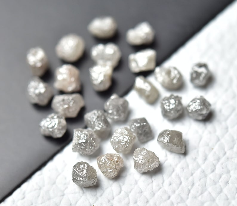 Puede incluir: Una colecci&oacute;n de diamantes grises en bruto y sin pulir. Los diamantes son de varios tama&ntilde;os y formas, y est&aacute;n esparcidos sobre una superficie blanca.