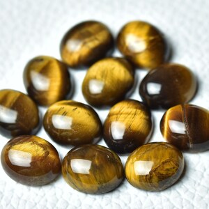 K&ouml;nnte beinhalten: Eine Sammlung von zehn ovalen Tigeraugen-Edelsteinen. Die Steine sind eine Mischung aus goldbraunen und dunkelbraunen Farben mit einem Chatoyance-Effekt.