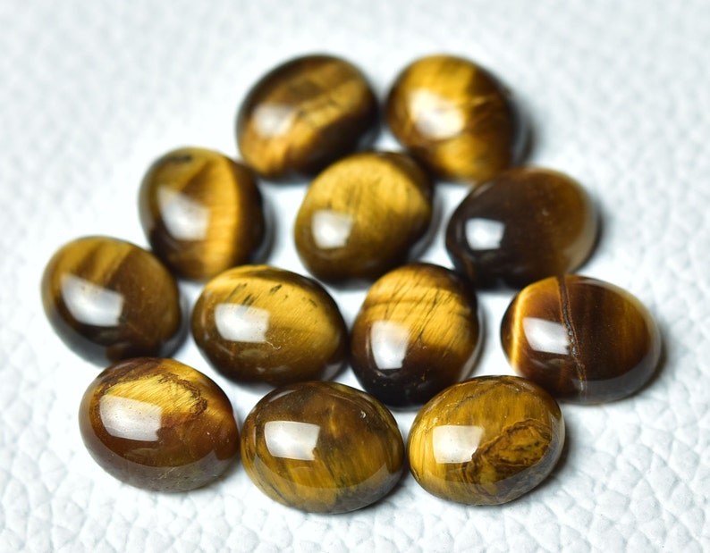 K&ouml;nnte beinhalten: Eine Sammlung von zehn ovalen Tigeraugen-Edelsteinen. Die Steine sind eine Mischung aus goldbraunen und dunkelbraunen Farben mit einem Chatoyance-Effekt.