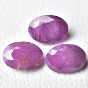 May include: Three oval-shaped, faceted gemstones with a purple hue. The gemstones are arranged in a row on a white surface.