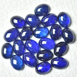 May include: A collection of oval-shaped, deep blue gemstones. The polished stones have a glossy finish, reflecting light. The gems are scattered on a white surface, showcasing their rich color and smooth texture. These could be used for jewelry making.