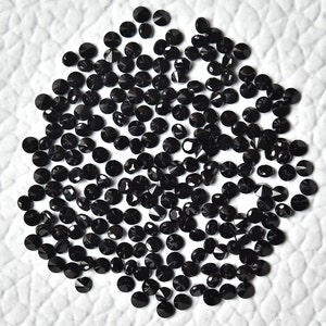 May include: A large quantity of black round gemstones, likely diamonds, scattered on a white surface. The gemstones are faceted and have a shiny, reflective surface.
