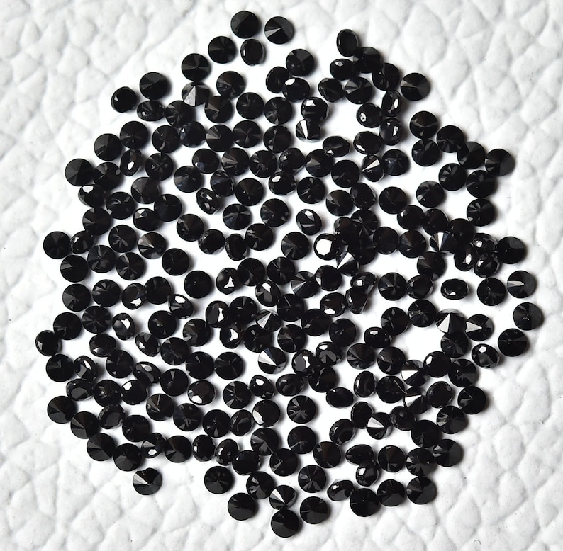 May include: A large quantity of black round gemstones, likely diamonds, scattered on a white surface. The gemstones are faceted and have a shiny, reflective surface.