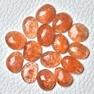 May include: A collection of 14 sunstone gemstones, each with a unique, faceted shape and a shimmering, orange-red colour. The stones are arranged in a circle on a white surface.