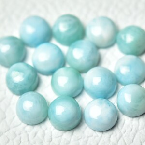 Puede incluir: Una colecci&oacute;n de 15 piedras preciosas cabuj&oacute;n redondas y lisas de color azul claro. Los cabujones est&aacute;n dispuestos en un c&iacute;rculo suelto sobre una superficie blanca.