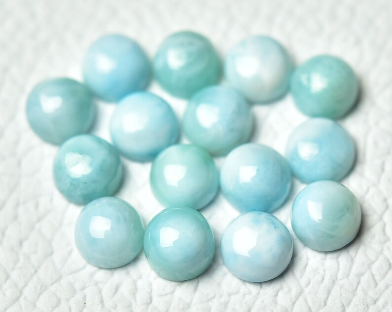 Puede incluir: Una colecci&oacute;n de 15 piedras preciosas cabuj&oacute;n redondas y lisas de color azul claro. Los cabujones est&aacute;n dispuestos en un c&iacute;rculo suelto sobre una superficie blanca.