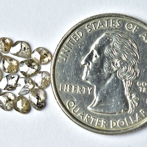 May include: A group of 15 small, rough, faceted, grey and white diamonds, with a hint of yellow, are scattered on a white surface. A US quarter is included for size comparison.