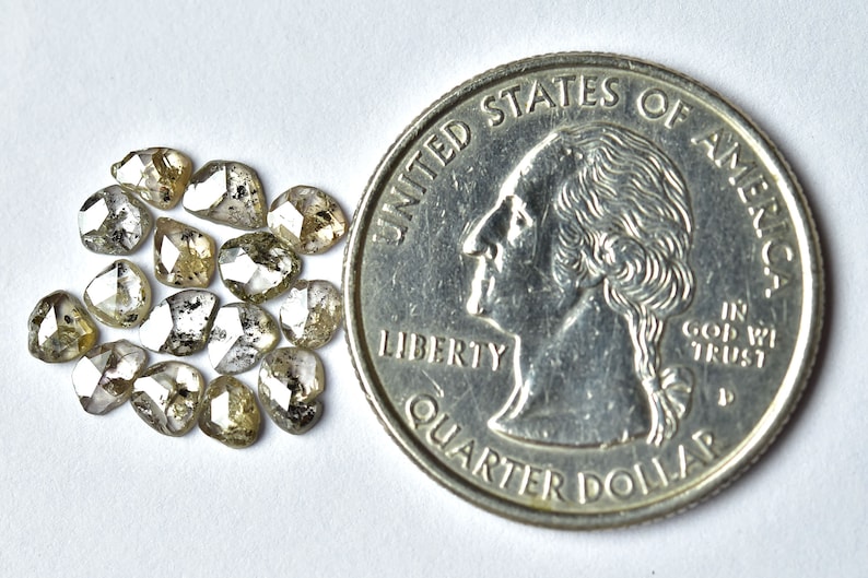 May include: A group of 15 small, rough, faceted, grey and white diamonds, with a hint of yellow, are scattered on a white surface. A US quarter is included for size comparison.