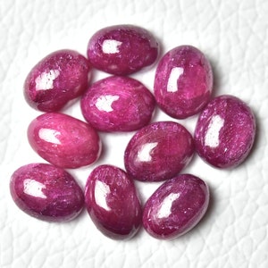 Może przedstawiać: Dziesięć owalnych, wypolerowanych rubinów w różnych odcieniach różu. Kamienie są ułożone w luźnym kole na białym tle.