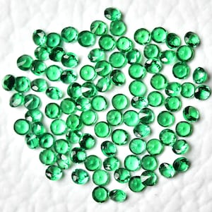 May include: A collection of small, round, faceted green gemstones. The gems are scattered across a white surface, each reflecting light with a brilliant sparkle. The stones vary slightly in size and shade, creating a visually appealing display of color and light.
