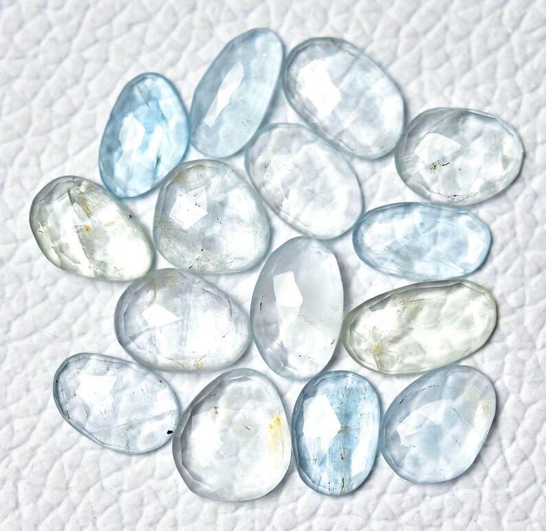 Puede incluir: Una colecci&oacute;n de 15 piedras preciosas de aguamarina azul claro, cada una con una forma ovalada facetada &uacute;nica. Las piedras est&aacute;n dispuestas sobre una superficie blanca.