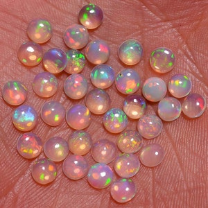 May include: A collection of round, iridescent opal gemstones in various shades of pink, white, and blue. The opals are arranged in a loose pile on a textured surface.