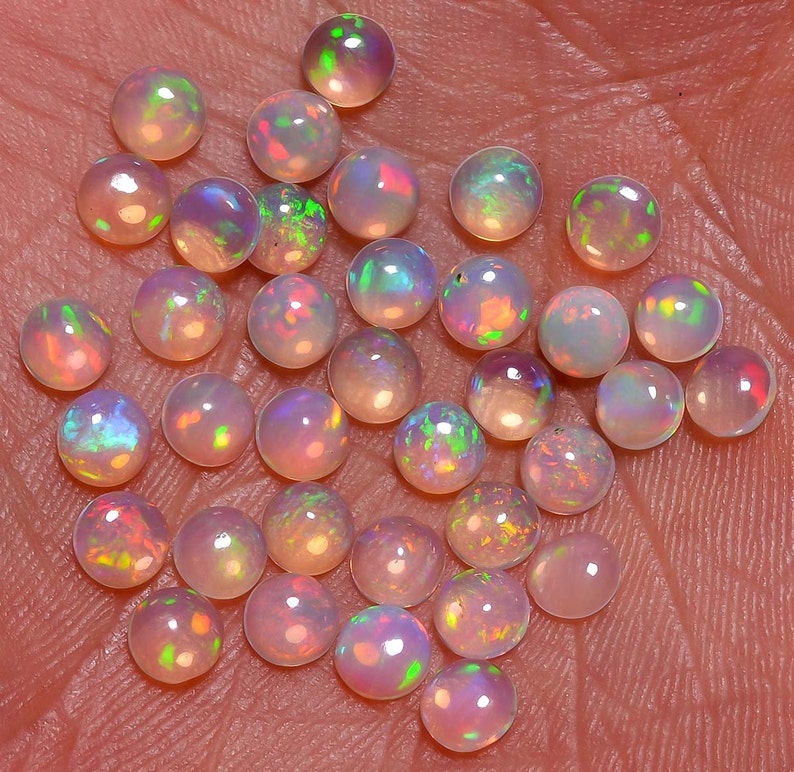 May include: A collection of round, iridescent opal gemstones in various shades of pink, white, and blue. The opals are arranged in a loose pile on a textured surface.