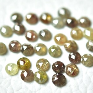 Op de afbeelding: Een verzameling ronde, gefacetteerde, ruwe diamanten in verschillende tinten bruin, groen en geel. De diamanten zijn verspreid over een wit oppervlak.