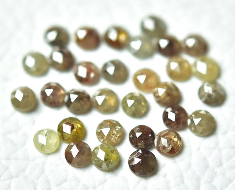 Op de afbeelding: Een verzameling ronde, gefacetteerde, ruwe diamanten in verschillende tinten bruin, groen en geel. De diamanten zijn verspreid over een wit oppervlak.