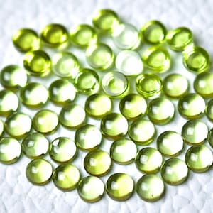 May include: A collection of round, green peridot gemstones, each with a smooth, polished surface. The stones are arranged in a circular pattern on a white background.