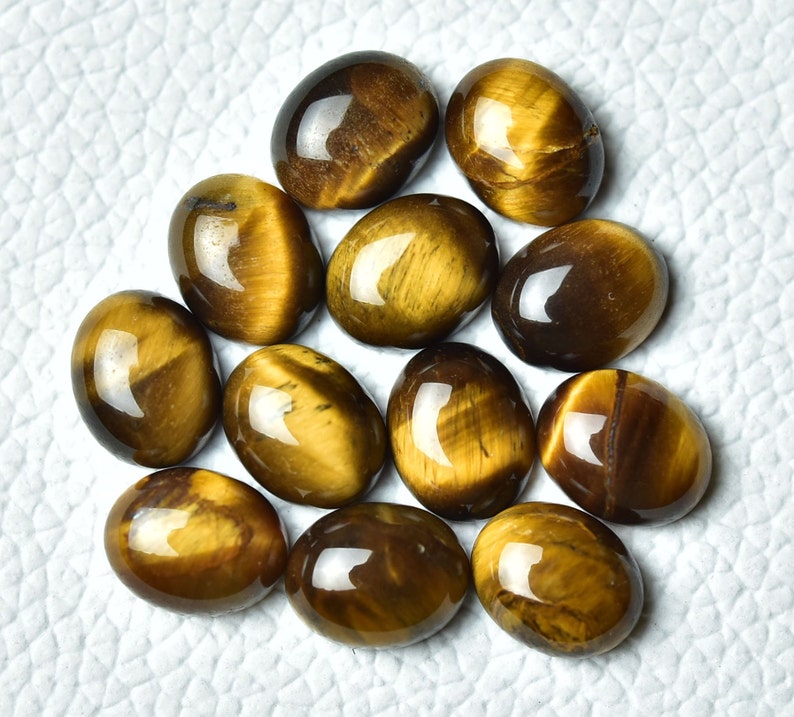 K&ouml;nnte beinhalten: Eine Sammlung von 12 ovalen Tigeraugen-Edelsteinen. Die Steine sind eine Mischung aus braunen und goldenen Farben mit einem chatoyanten Effekt.