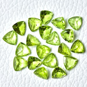 May include: A collection of 18 triangular green peridot gemstones, each with a unique cut and facet pattern. The stones are arranged on a white surface.