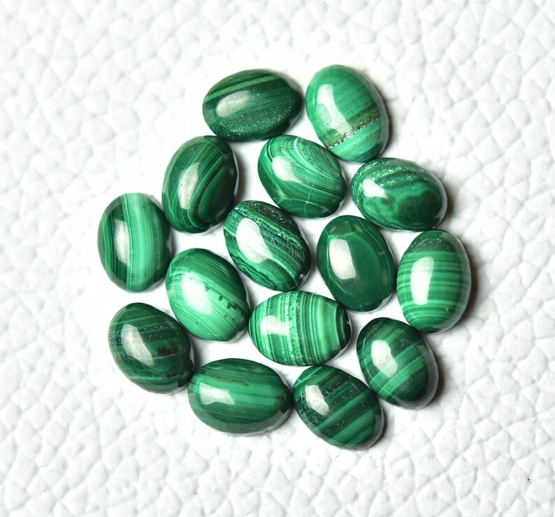 May include: A collection of 15 oval-shaped malachite gemstones. The stones are a deep green color with intricate patterns of lighter and darker green.