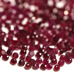 May include: A close-up of a pile of small, round, red gemstones. The gemstones are faceted and have a shiny, polished finish.