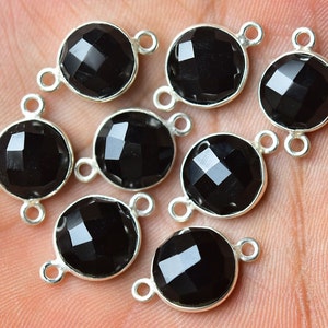 Pode incluir: Close-up de oito pingentes redondos de pedras preciosas pretas facetadas com engastes de metal prateado. Cada pingente tem dois pequenos laços para fixar em joias. As pedras têm uma superfície polida e reflexiva.