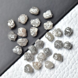 Puede incluir: Una colecci&oacute;n de diamantes grises en bruto, tambi&eacute;n conocidos como diamantes en bruto, en varios tama&ntilde;os y formas. Los diamantes est&aacute;n dispersos sobre una superficie blanca.