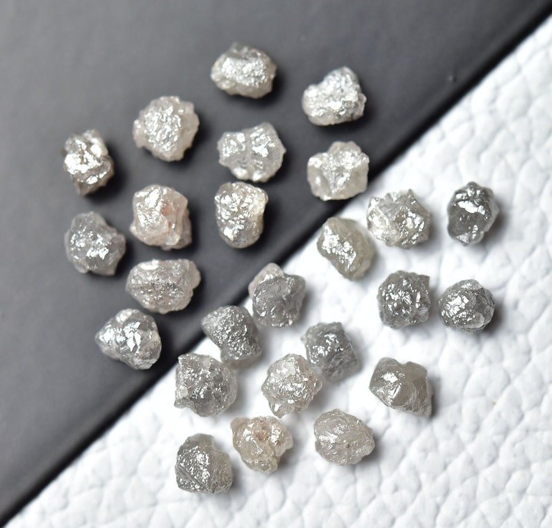 Puede incluir: Una colecci&oacute;n de diamantes grises en bruto, tambi&eacute;n conocidos como diamantes en bruto, en varios tama&ntilde;os y formas. Los diamantes est&aacute;n dispersos sobre una superficie blanca.