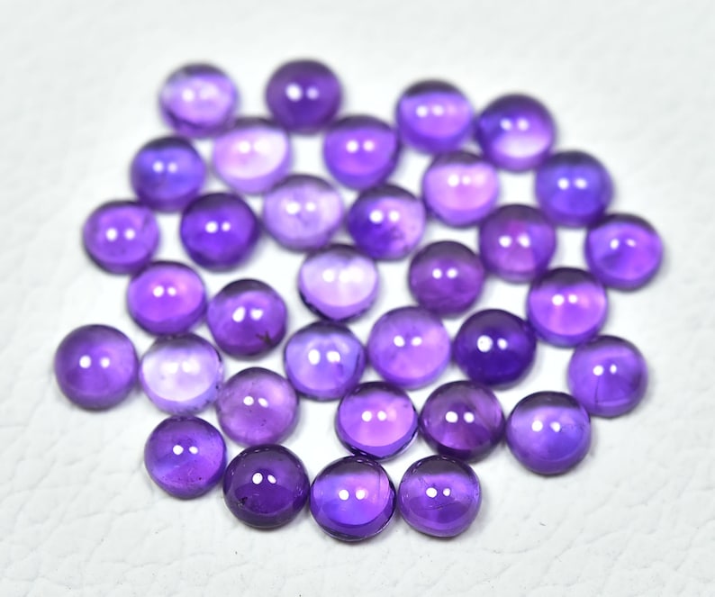 May include: A collection of 25 round, purple amethyst cabochons. The cabochons are smooth and polished, with a slight variation in color and size.