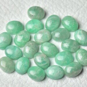 Peut inclure: Une collection de 20 pierres pr&eacute;cieuses vertes de forme ovale, probablement de l'amazonite, avec une finition lisse et polie. Les pierres sont dispos&eacute;es de mani&egrave;re al&eacute;atoire sur une surface blanche.